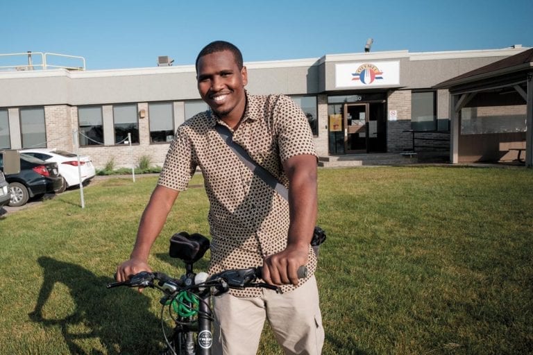 Image de Faram Hassan Doudoub, souriant avec son vélo devant l'usine Olymel