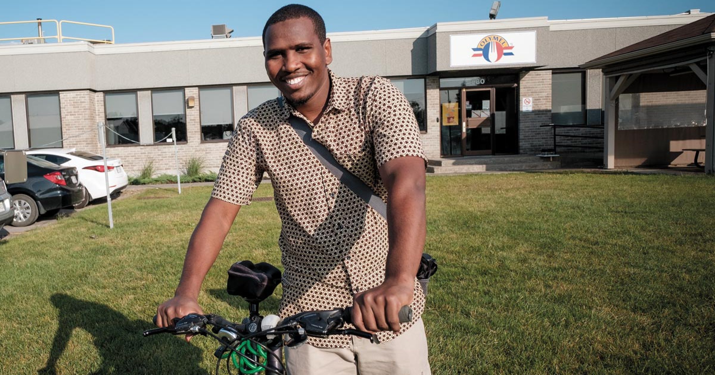 Image de Faram Hassan Doudoub, souriant avec son vélo devant l'usine Olymel