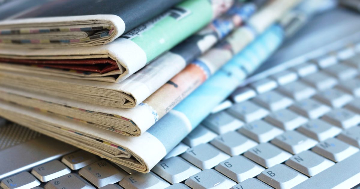 Newspapers and laptop. Pile of daily papers with news on the computer. Pages with headlines, articles folded and stacked on keypad of electronic device. Modern gadget and old journals, focus on paper