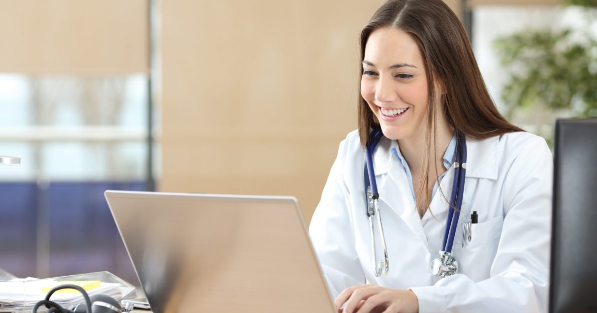 Happy doctor using a laptop writing content at consultation